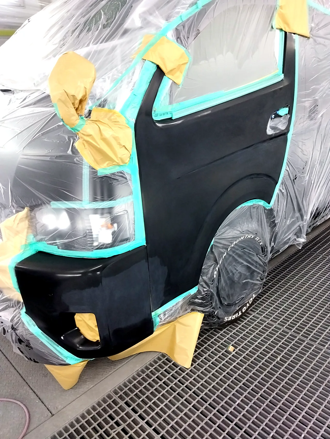 塗装ブースでビニールに覆われたハイエース。バンパー左角と左フロントドア1枚を出したところ(クリヤーの前)