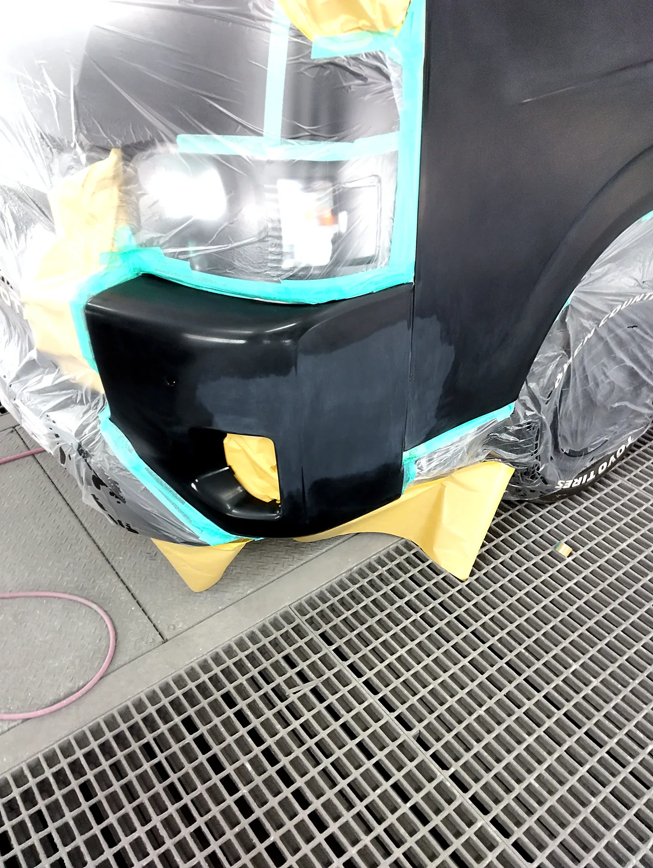 塗装ブースのハイエースのバンパー左角。ヘッドライトの縁にテープ、フォグランプの穴は裏から紙(クリヤーの前)