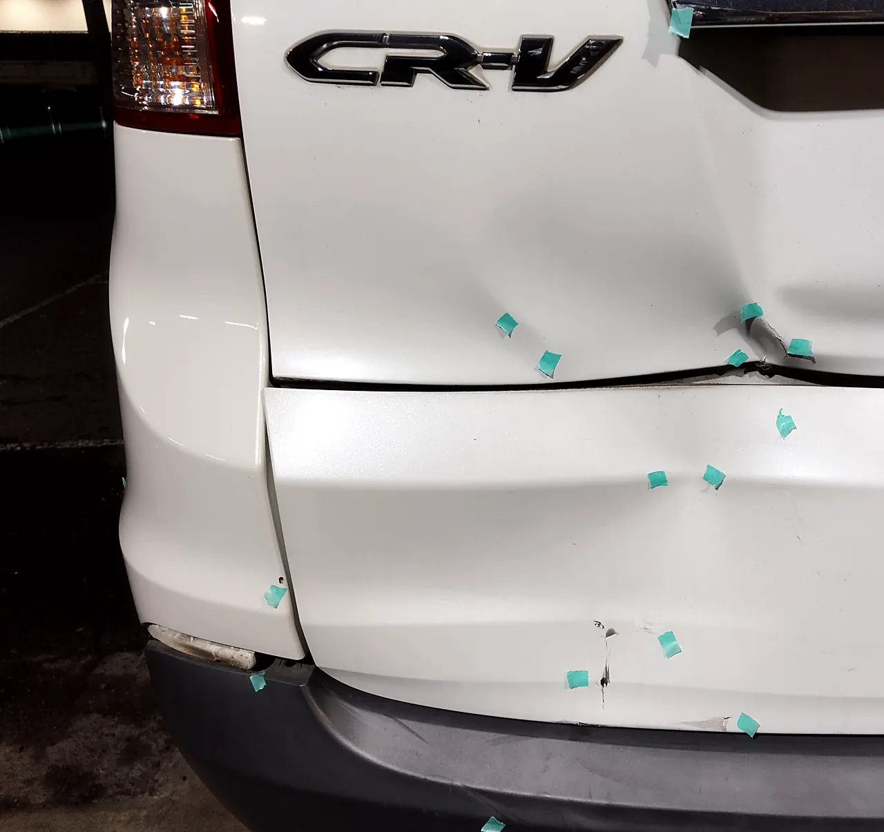 修理前のホンダ CR-V のエンブレムの下。バックドアの下側と、その下の白い板がへこみ、緑のテープの印
