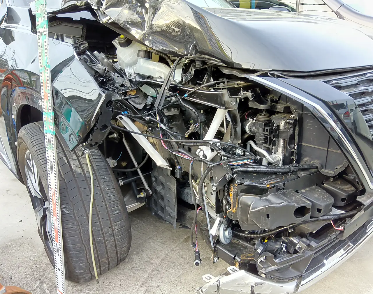 エクストレイル（T33）の右前の寄り。ヘッドランプがなく、配線や中の部品がむき出しで、右前輪が押し込まれている（修理前・目盛りの棒）